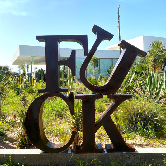 Sculpture typographique "FUCK" en métal oxydé posée dans un jardin, lettres découpées à la main avec patine naturelle
